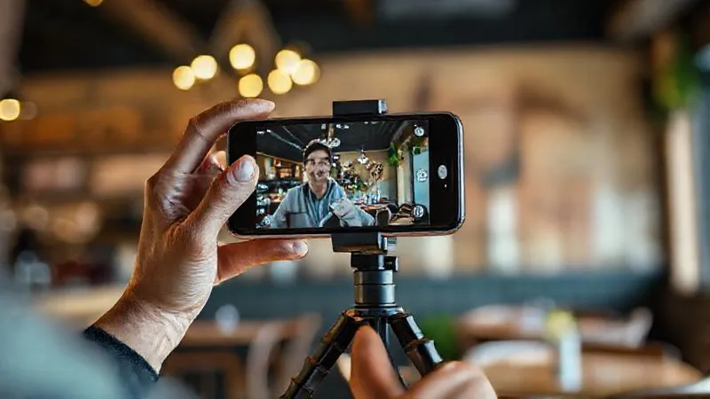 A photorealistic image of a person holding a modern smartphone horizontally, mounted on a small, flexible tripod, recording themselves in a well-lit cafe with a blurred background. Cinematic lighting, sharp focus on the smartphone and person, depth of field, 8K hyper-detailed, shot on a high-end DSLR.