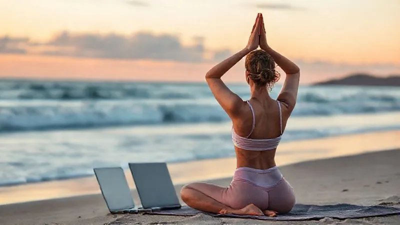 A photorealistic image of a digital nomad (gender-neutral) doing yoga poses on a beach at sunrise, with a laptop closed beside them. The scene exudes tranquility and balance. Cinematic lighting casts warm hues, with sharp focus on the individual and a soft depth of field blurring the ocean waves. Shot on a high-end DSLR, 8K hyper-detailed.