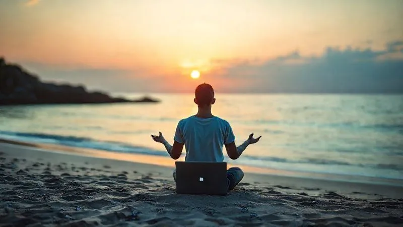 A photorealistic image of a digital nomad meditating peacefully on a tranquil beach at sunrise, with a laptop subtly placed in the background. The scene exudes calm, focus, and connection with nature. Cinematic lighting, sharp focus on the meditating figure, depth of field blurring the background, 8K hyper-detailed, professional photography, shot on a high-end DSLR.