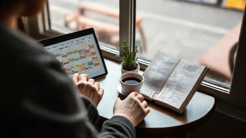 A photorealistic image of a digital nomad sitting by a window in a cozy cafe, reviewing a digital calendar and a physical journal. The calendar shows color-coded tasks and flexible deadlines, while the journal has notes on weekly progress and adaptations. A cup of coffee and a small plant are on the table. Soft, natural light, sharp focus on the hands and journal, depth of field blurring the cafe background, 8K hyper-detailed, shot on a high-end DSLR.