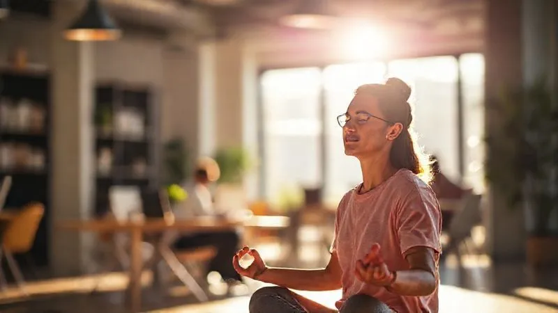 A photorealistic image of a person meditating calmly in a vibrant, sunlit co-working space, surrounded by subtle, blurred activity. The focus is on their serene expression and posture, suggesting inner peace and concentration amidst potential distractions. Cinematic lighting, 8K, sharp focus on the person, depth of field blurring the background, shot on a high-end DSLR.