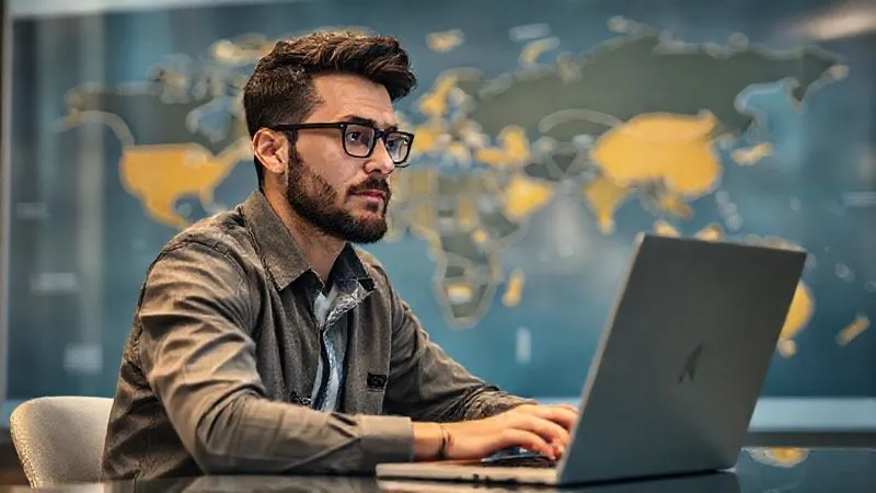 A photorealistic image of a digital nomad sitting at a laptop, looking confident and thoughtful, with a world map subtly blurred in the background, representing global market value research and personal financial planning. Professional photography, 8K, cinematic lighting, sharp focus, depth of field.