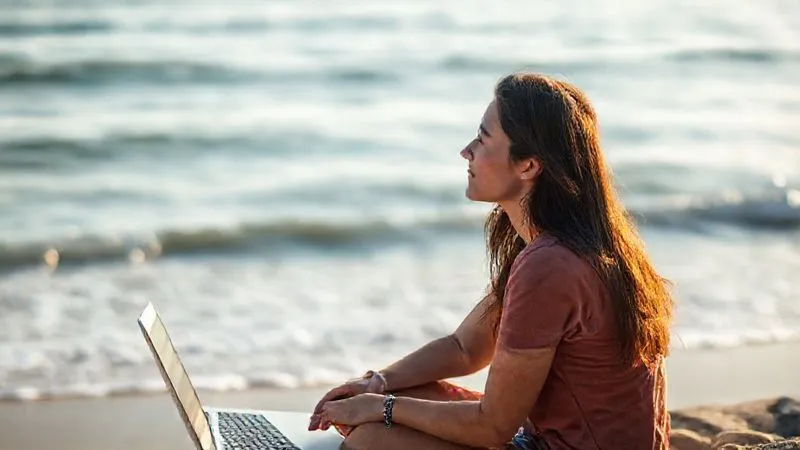 A photorealistic, professional photography of a digital nomad, mid-20s, sitting by a serene beach, laptop closed beside them. They are looking out at the ocean with a peaceful, reflective expression, holding a seashell. The scene conveys a sense of calm, disconnection, and mental recharge. Soft morning light, sharp focus on the nomad, depth of field blurring the waves, 8K, shot on a high-end DSLR.