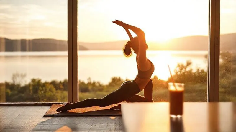 A photorealistic image of a digital nomad doing morning stretches on a yoga mat by a large window overlooking a serene lake at sunrise, a healthy breakfast smoothie on a nearby table. Soft, natural cinematic lighting, sharp focus on the nomad, depth of field blurring the background, 8K hyper-detailed.