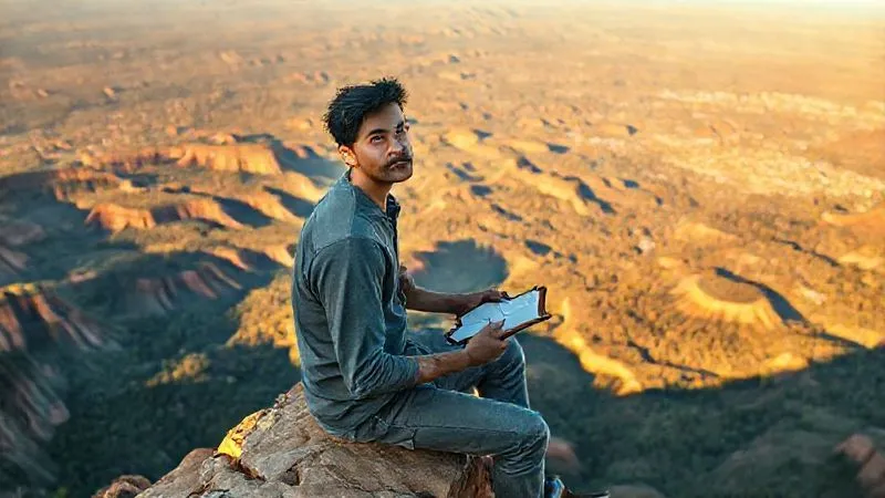 A photorealistic image of a digital nomad sitting on a cliff edge overlooking a vast, intricate landscape (e.g., a canyon, a dense forest, or a complex city skyline), holding a small, open notebook and a pen, with a look of deep contemplation and discovery. The lighting is golden hour, casting long, dramatic shadows. Sharp focus on the nomad's thoughtful expression and the notebook, with the breathtaking landscape blurring into the depth of field. 8K hyper-detailed, professional photography, shot on a high-end DSLR.