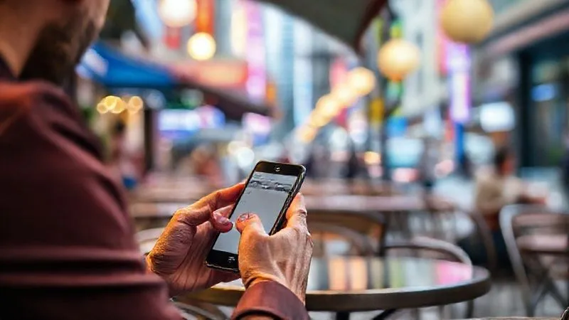 A photorealistic, professional photography, 8K, cinematic lighting, sharp focus, depth of field, shot on a high-end DSLR, showing a digital nomad sitting casually in a vibrant, outdoor cafe in a bustling foreign city. The person is holding a smartphone, actively typing notes into the Notion app. The screen is clearly visible showing a minimalist note-taking interface. The background is slightly blurred, capturing the lively atmosphere without distracting from the focused action of capturing information.