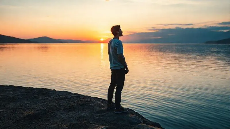 A photorealistic image of a lone digital nomad standing at the edge of a vast, tranquil lake at sunrise, looking out at the horizon with a sense of calm determination. The metaphor is about overcoming the overwhelming 'ocean' of information and finding clarity. Cinematic lighting, professional photography, 8K, sharp focus on the nomad's thoughtful expression, depth of field blurring the serene landscape. Shot on a high-end DSLR.