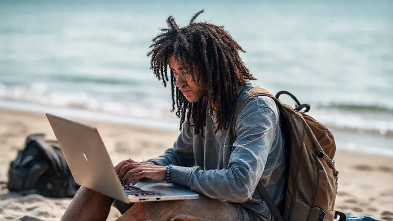 A photorealistic image of a young, diverse digital nomad working diligently on a laptop on a beach, with a backpack nearby and a calm ocean in the background. The lighting is soft and natural, emphasizing focus despite the serene environment. 8K hyper-detailed, shot on a high-end DSLR with depth of field.